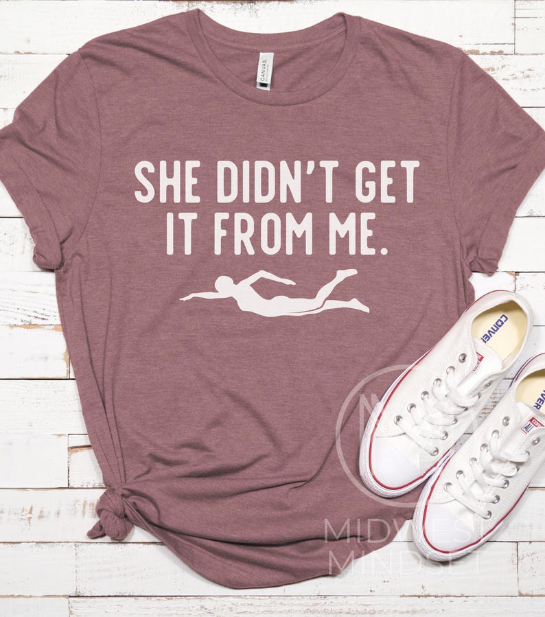 May include: A mauve t-shirt with the text "She didn't get it from me." in white. A white silhouette of a swimmer is below the text.