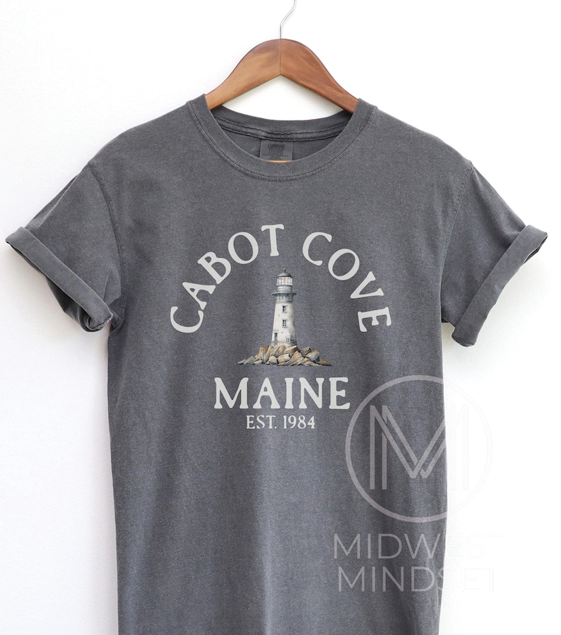 以下が含まれることがあります： 袖をまくった濃いグレーのTシャツ。岩の上に灯台の白いグラフィックがあり、その上下に「CABOT COVE MAINE EST. 1984」の文字がアーチ状に配置されています。