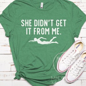 May include: Green t-shirt with the text "She didn't get it from me." and a white silhouette of a swimmer.