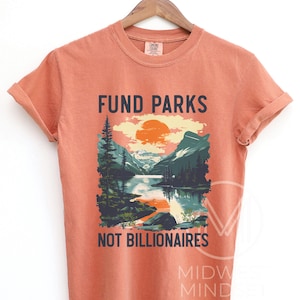 May include: A burnt orange t-shirt with a graphic of a mountain lake scene and the text "Fund Parks Not Billionaires".