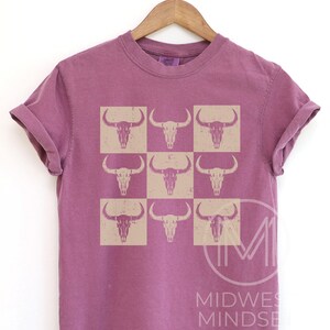 Pu&ograve; includere: Una t-shirt color malva con un motivo di nove teschi di toro beige disposti in una griglia.
