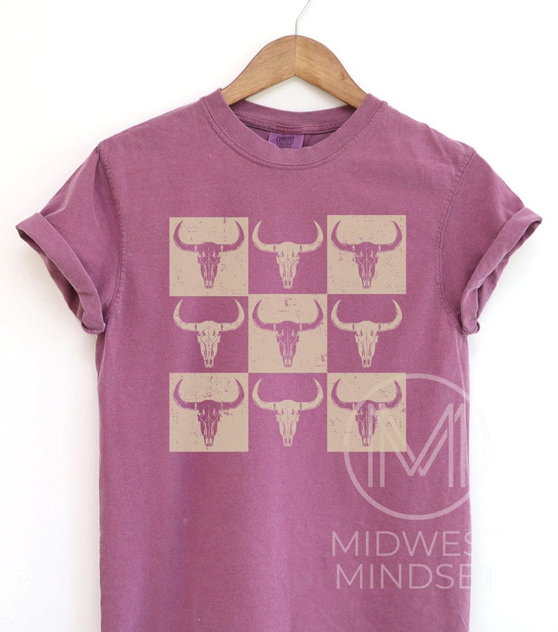 Pu&ograve; includere: Una t-shirt color malva con un motivo di nove teschi di toro beige disposti in una griglia.