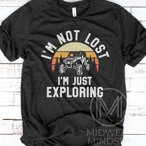 K&ouml;nnte beinhalten: Ein dunkelgraues T-Shirt mit einer Vintage-Sonnenuntergangsgrafik, die eine Silhouette eines vierr&auml;drigen Gel&auml;ndewagens zeigt. Der Text "Ich bin nicht verloren, ich erkunde nur" ist in Wei&szlig; auf das Shirt gedruckt.