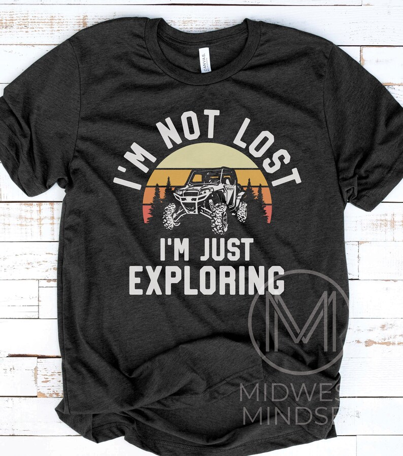 K&ouml;nnte beinhalten: Ein dunkelgraues T-Shirt mit einer Vintage-Sonnenuntergangsgrafik, die eine Silhouette eines vierr&auml;drigen Gel&auml;ndewagens zeigt. Der Text "Ich bin nicht verloren, ich erkunde nur" ist in Wei&szlig; auf das Shirt gedruckt.
