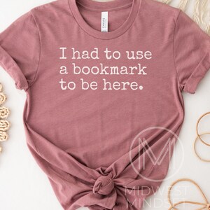 May include: A dusty rose t-shirt with the text "I had to use a bookmark to be here" in white. The shirt is knotted at the bottom and has a crew neck. The brand tag is visible at the neckline. The shirt is a casual wear item.