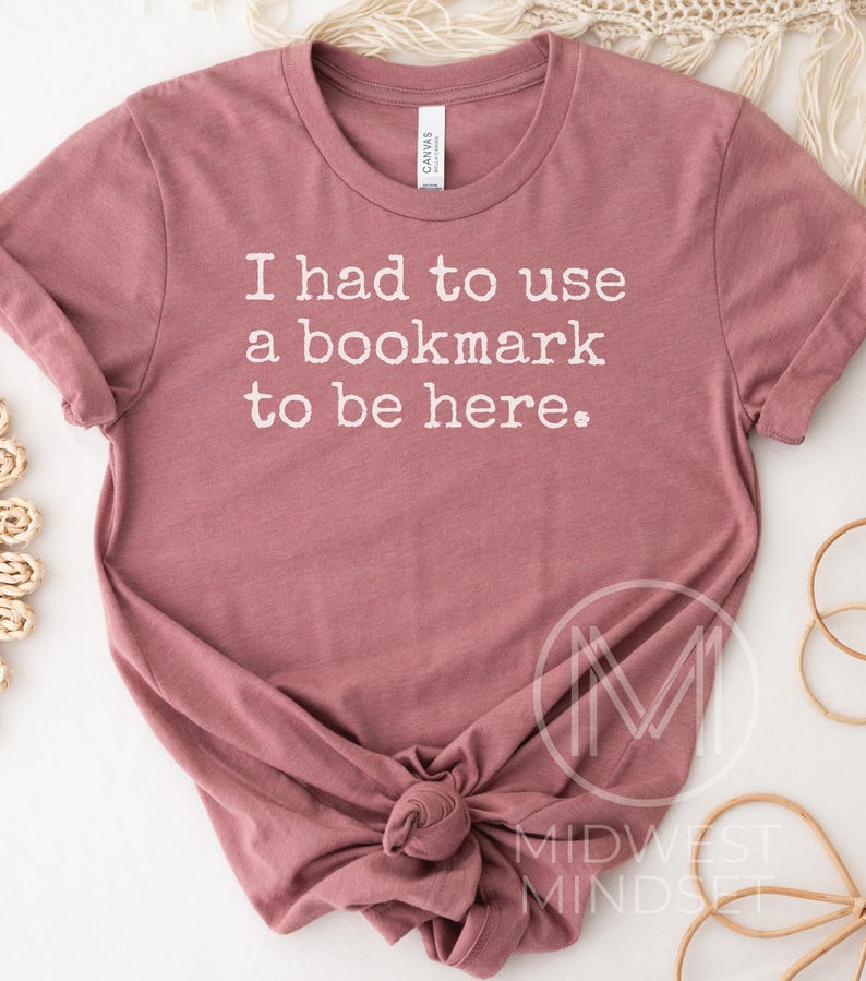 May include: A dusty rose t-shirt with the text "I had to use a bookmark to be here" in white. The shirt is knotted at the bottom and has a crew neck. The brand tag is visible at the neckline. The shirt is a casual wear item.