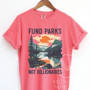 May include: A pink t-shirt with a graphic design of a mountain landscape with a lake and the text "Fund Parks Not Billionaires".