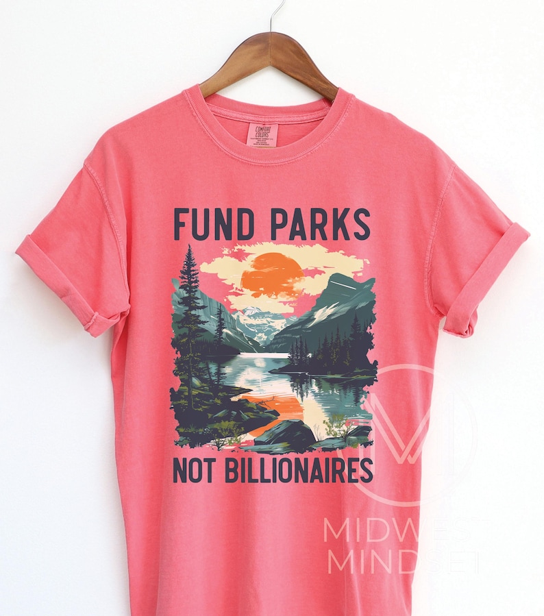 May include: A pink t-shirt with a graphic design of a mountain landscape with a lake and the text "Fund Parks Not Billionaires".