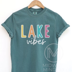 Puede incluir: Camiseta de color verde azulado con las palabras "LAKE vibes" en letras de bloque coloridas. La palabra "LAKE" est&aacute; en amarillo, naranja, turquesa y rosa. La palabra "vibes" est&aacute; en cursiva. La camiseta est&aacute; colgada en una percha de madera.