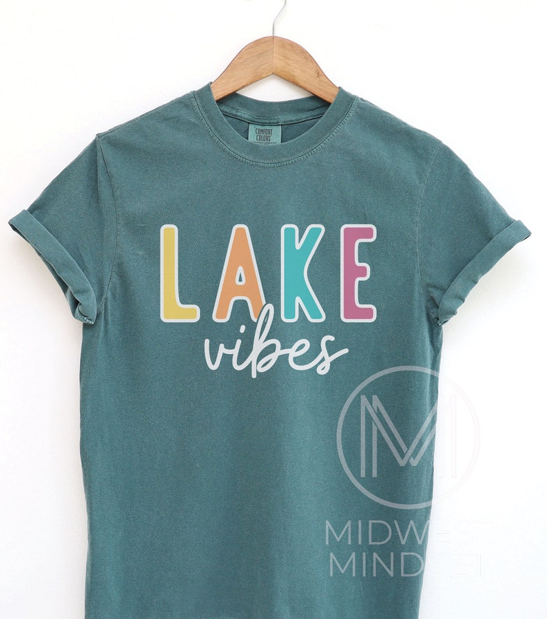 Puede incluir: Camiseta de color verde azulado con las palabras "LAKE vibes" en letras de bloque coloridas. La palabra "LAKE" est&aacute; en amarillo, naranja, turquesa y rosa. La palabra "vibes" est&aacute; en cursiva. La camiseta est&aacute; colgada en una percha de madera.