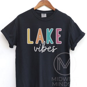 Puede incluir: Camiseta negra con las palabras "LAKE vibes" en letras coloridas. La palabra "LAKE" es amarilla, naranja, turquesa y rosa. La palabra "vibes" est&aacute; en cursiva. La camiseta est&aacute; colgada en una percha de madera.
