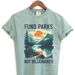 May include: A light green t-shirt with a graphic design of a mountain landscape with a lake and a sunset. The text "Fund Parks Not Billionaires" is printed on the shirt.