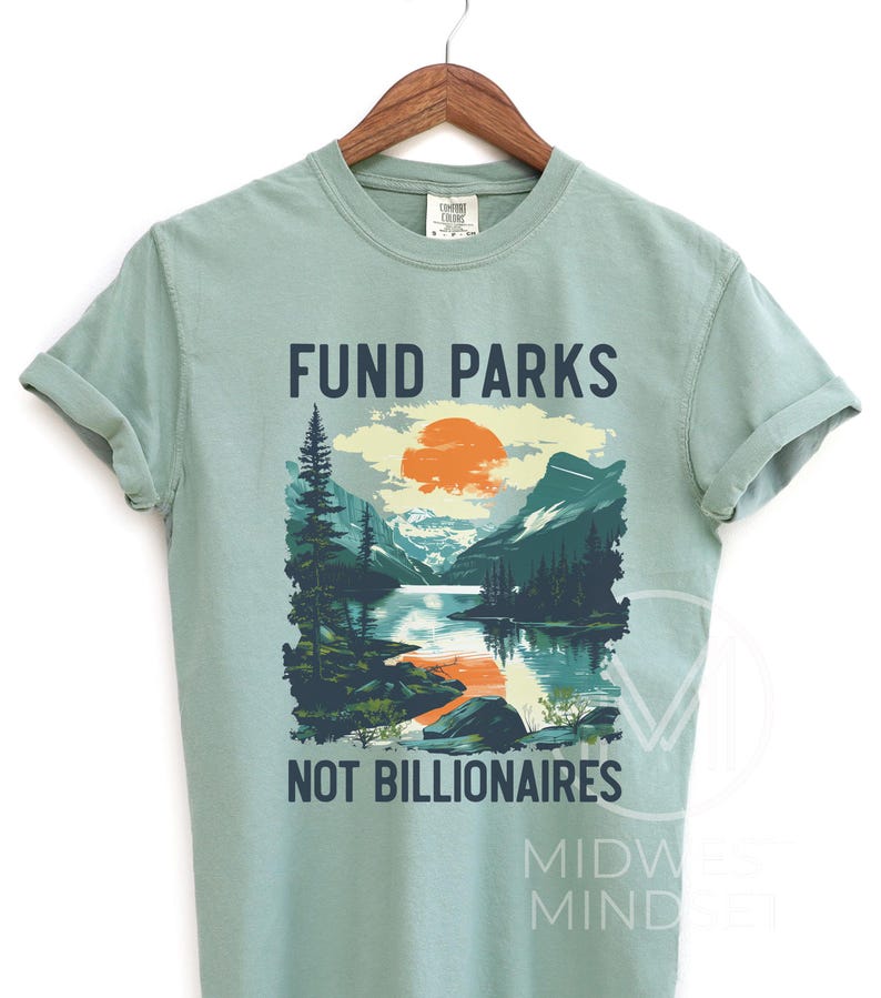 May include: A light green t-shirt with a graphic design of a mountain landscape with a lake and a sunset. The text "Fund Parks Not Billionaires" is printed on the shirt.