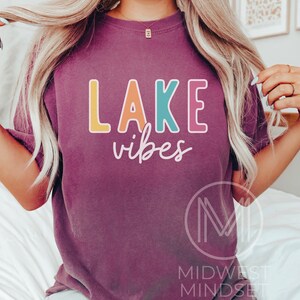 Puede incluir: Una camiseta morada con las palabras "LAKE vibes" en letras de bloque coloridas. La palabra "LAKE" est&aacute; en amarillo, turquesa, rosa y naranja. La palabra "vibes" est&aacute; en una fuente cursiva.