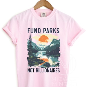 May include: Pink t-shirt with a graphic of a mountain landscape and a sunset. The text "Fund Parks Not Billionaires" is printed on the shirt.