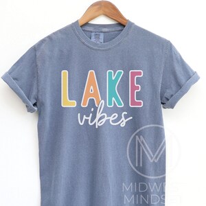 Puede incluir: Una camiseta azul gris&aacute;ceo con las palabras "LAKE vibes" impresas en la parte delantera en letras coloridas. La palabra "LAKE" est&aacute; en amarillo, naranja, turquesa y rosa. La camiseta est&aacute; colgada en una percha de madera.