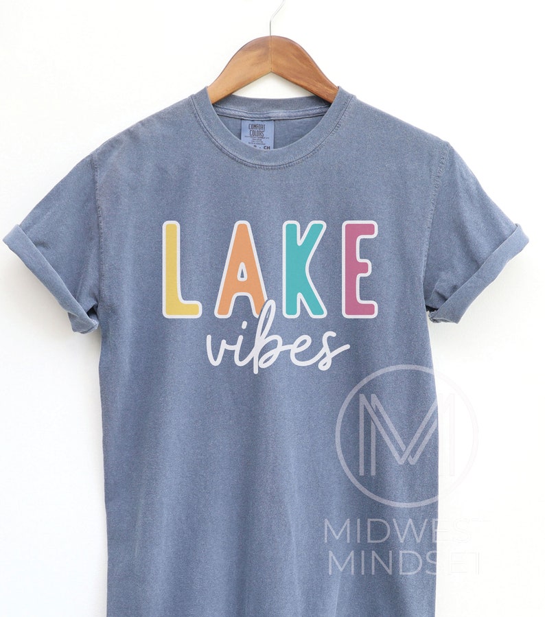 Puede incluir: Una camiseta azul gris&aacute;ceo con las palabras "LAKE vibes" impresas en la parte delantera en letras coloridas. La palabra "LAKE" est&aacute; en amarillo, naranja, turquesa y rosa. La camiseta est&aacute; colgada en una percha de madera.