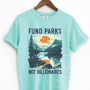 May include: A light blue t-shirt with a graphic design of a mountain scene with a lake and a setting sun. The text "Fund Parks Not Billionaires" is printed on the shirt.