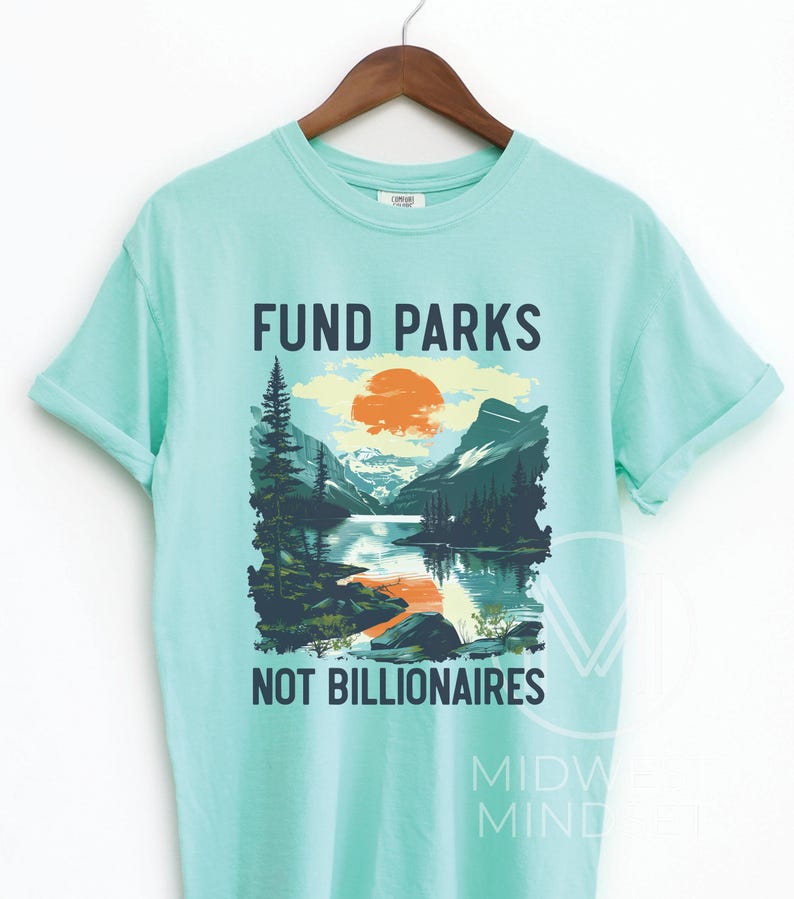 May include: A light blue t-shirt with a graphic design of a mountain scene with a lake and a setting sun. The text "Fund Parks Not Billionaires" is printed on the shirt.