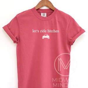 Puede incluir: Una camiseta roja con el texto "let's ride bitches" y un peque&ntilde;o gr&aacute;fico de coche en blanco. La camiseta est&aacute; colgada en una percha de madera. La camiseta es de color liso y tiene mangas cortas.