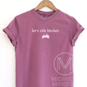 Puede incluir: Camiseta malva con mangas dobladas. La camiseta presenta el texto "let's ride bitches" sobre un peque&ntilde;o gr&aacute;fico de coche blanco. La camiseta est&aacute; colgada de una percha de madera.