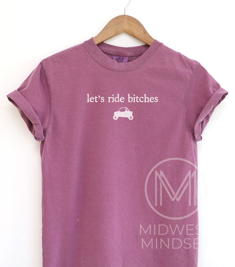 Puede incluir: Camiseta malva con mangas dobladas. La camiseta presenta el texto "let's ride bitches" sobre un peque&ntilde;o gr&aacute;fico de coche blanco. La camiseta est&aacute; colgada de una percha de madera.
