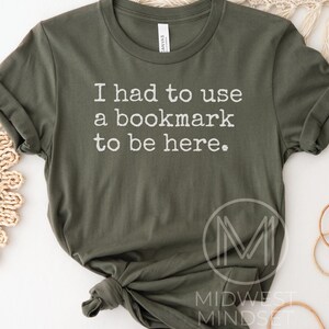 May include: Olive green t-shirt with the text "I had to use a bookmark to be here." The shirt has short sleeves and a crew neck. The text is in a simple, white font. The shirt is knotted at the bottom.