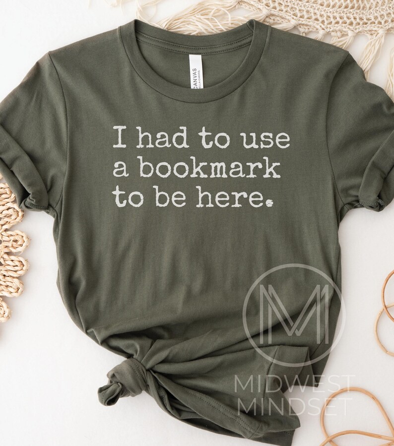 May include: Olive green t-shirt with the text "I had to use a bookmark to be here." The shirt has short sleeves and a crew neck. The text is in a simple, white font. The shirt is knotted at the bottom.