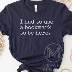 May include: Navy blue t-shirt with the text "I had to use a bookmark to be here." in white. The shirt also features a logo that says "Midwest Mindset."