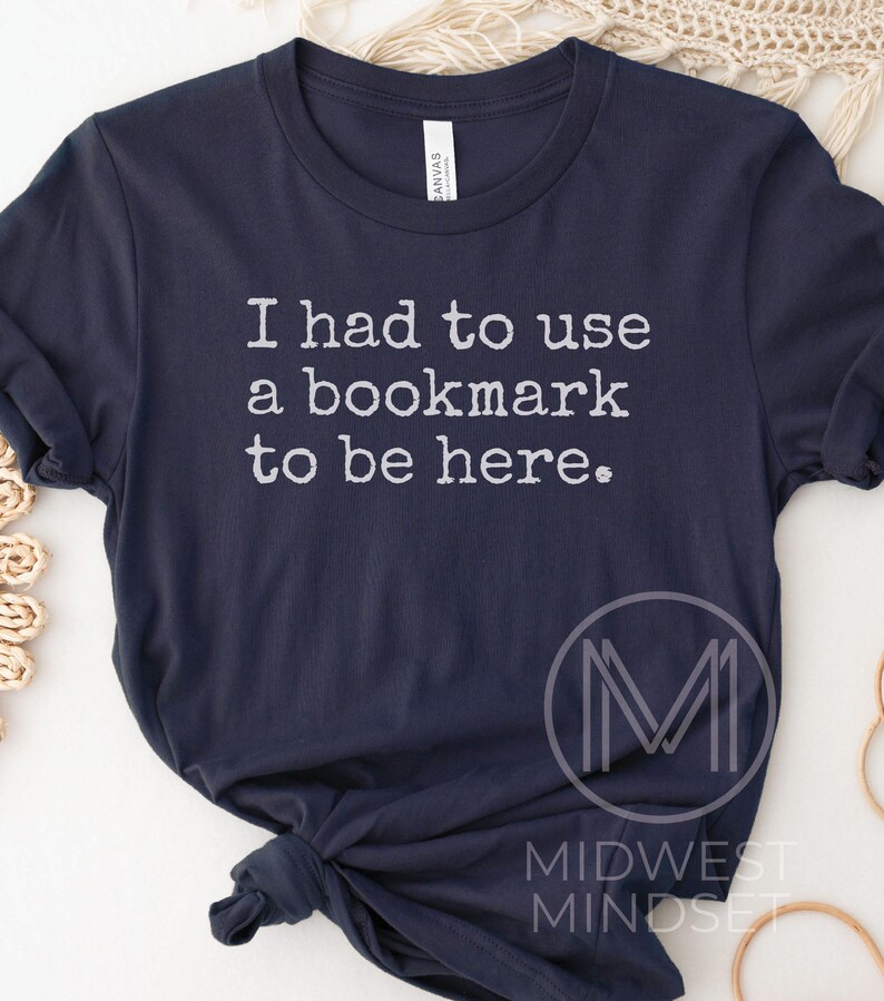 May include: Navy blue t-shirt with the text "I had to use a bookmark to be here." in white. The shirt also features a logo that says "Midwest Mindset."