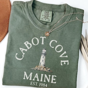 以下が含まれることがあります： 「Cabot Cove Maine」の文字と灯台のグラフィックがプリントされたオリーブグリーンのTシャツ。Tシャツには「EST. 1984」の文字も。金色のネックレスとペンダントがTシャツの上に置かれています。Comfort Colorsブランド。