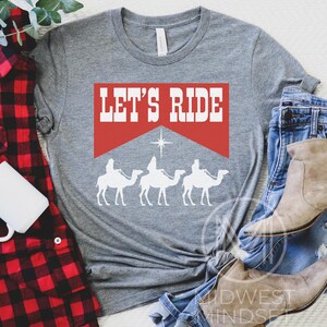 Pu&ograve; includere: Maglietta grigia con uno striscione rosso che dice "Let's Ride". Lo striscione &egrave; sopra una silhouette di tre re magi che cavalcano cammelli, con una stella sopra di loro.