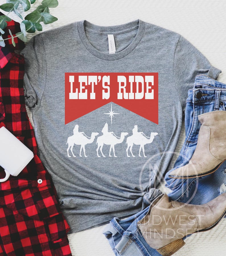 Pu&ograve; includere: Maglietta grigia con uno striscione rosso che dice "Let's Ride". Lo striscione &egrave; sopra una silhouette di tre re magi che cavalcano cammelli, con una stella sopra di loro.