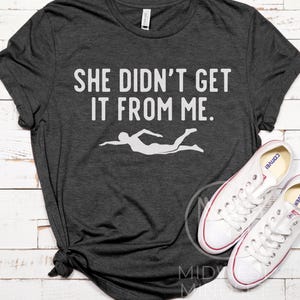 May include: A dark gray t-shirt with the text "She didn't get it from me." in white lettering. A white silhouette of a swimmer is below the text.