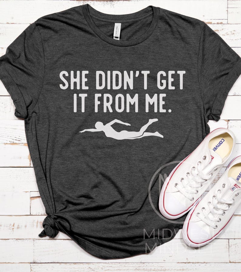 May include: A dark gray t-shirt with the text "She didn't get it from me." in white lettering. A white silhouette of a swimmer is below the text.