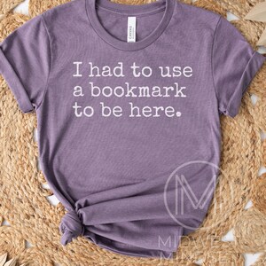 May include: A purple t-shirt with the text "I had to use a bookmark to be here." The shirt is made of soft material and has a crew neck. The sleeves are rolled up. The shirt is knotted at the bottom.