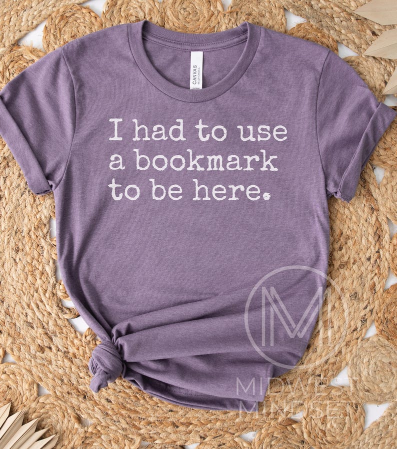May include: A purple t-shirt with the text "I had to use a bookmark to be here." The shirt is made of soft material and has a crew neck. The sleeves are rolled up. The shirt is knotted at the bottom.