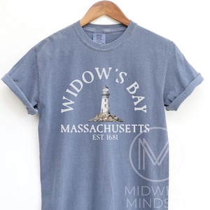 May include: A blue short-sleeve crew neck t-shirt featuring a graphic of a lighthouse on rocks. The text reads WIDOW'S BAY, MASSACHUSETTS, EST. 1681. The shirt is displayed on a wooden hanger, showcasing a casual style with rolled sleeves and a vintage look.