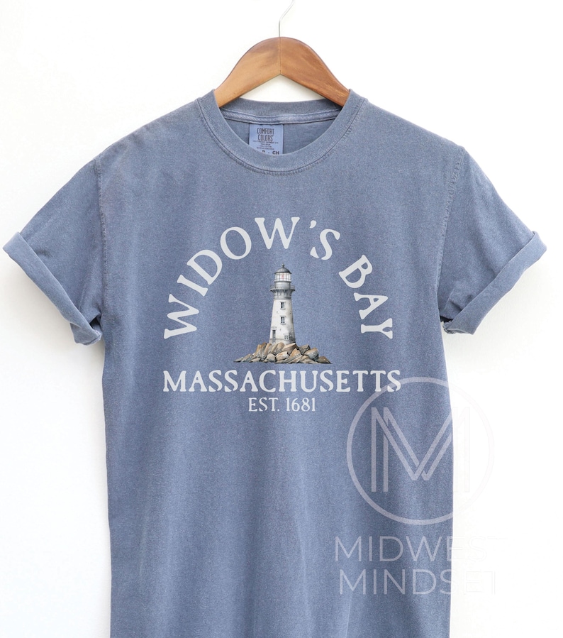 May include: A blue short-sleeve crew neck t-shirt featuring a graphic of a lighthouse on rocks. The text reads WIDOW'S BAY, MASSACHUSETTS, EST. 1681. The shirt is displayed on a wooden hanger, showcasing a casual style with rolled sleeves and a vintage look.