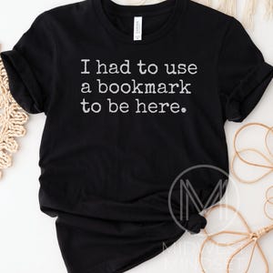 May include: Black t-shirt with the text "I had to use a bookmark to be here" in white. The shirt is made of a soft material and has rolled-up sleeves. The shirt is a casual wear item.