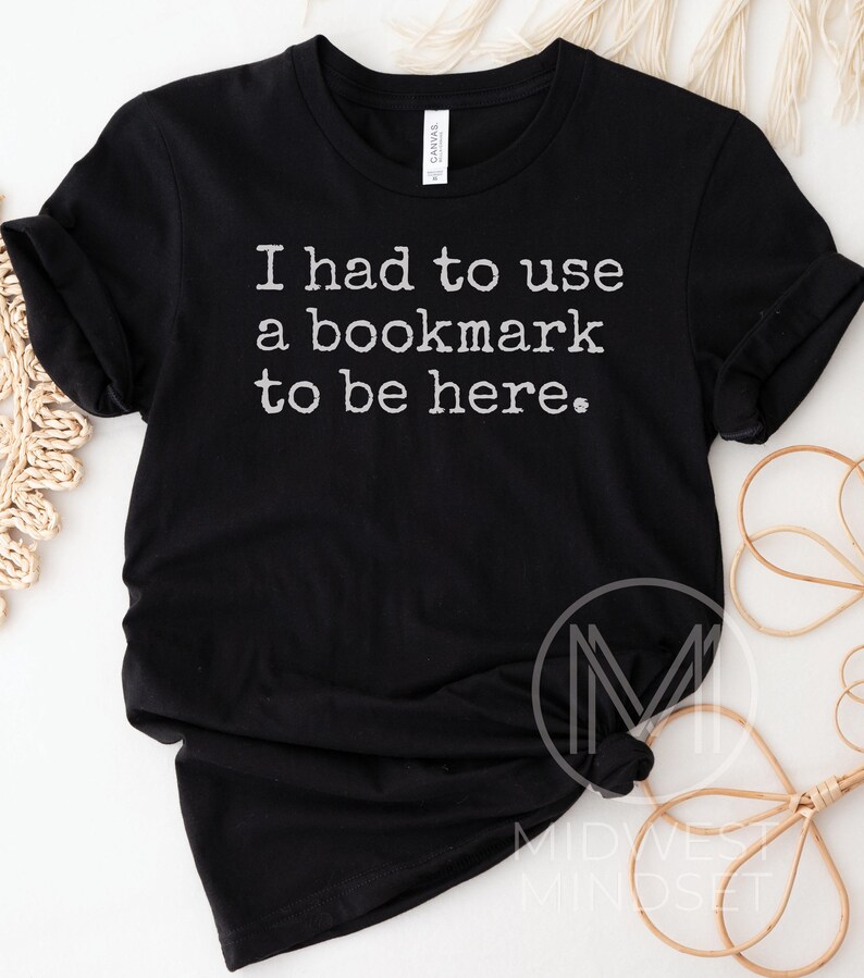 May include: Black t-shirt with the text "I had to use a bookmark to be here" in white. The shirt is made of a soft material and has rolled-up sleeves. The shirt is a casual wear item.