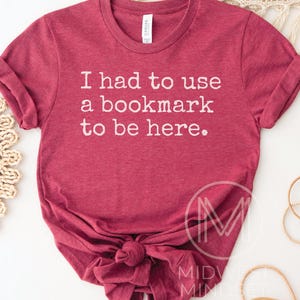 May include: A maroon t-shirt with the text "I had to use a bookmark to be here." The shirt is knotted at the bottom and has rolled-up sleeves. The shirt is made of a soft material.