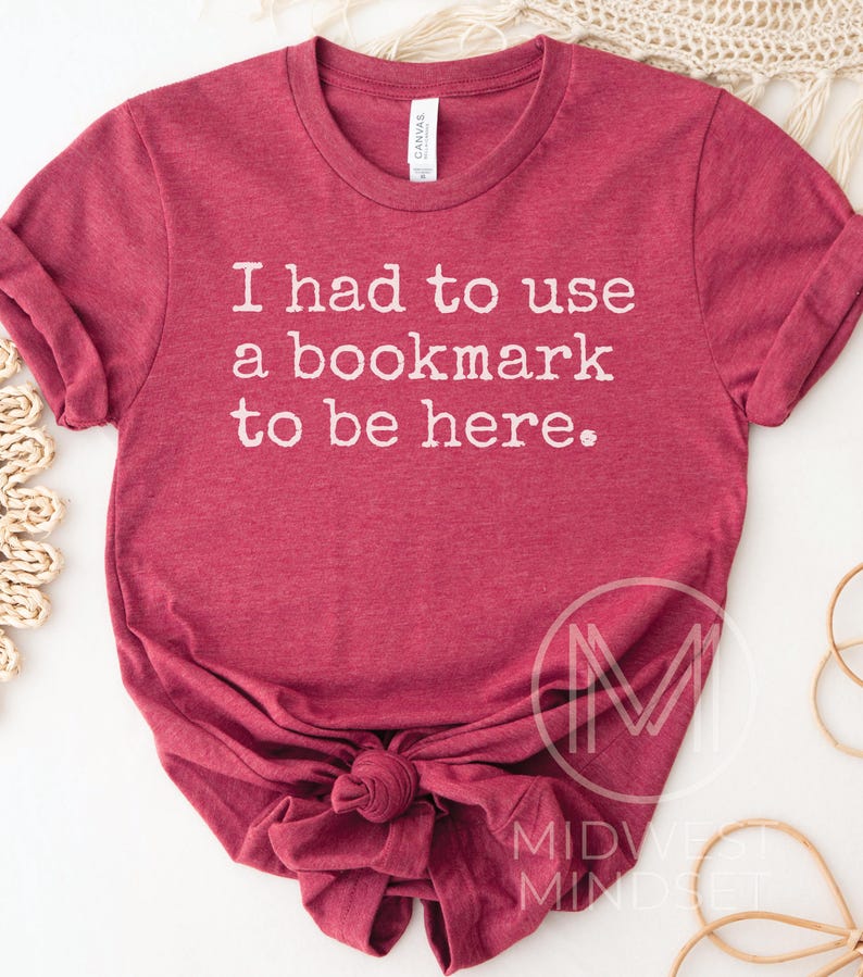May include: A maroon t-shirt with the text "I had to use a bookmark to be here." The shirt is knotted at the bottom and has rolled-up sleeves. The shirt is made of a soft material.