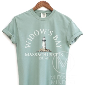 May include: A sage green short-sleeve t-shirt featuring a graphic of a lighthouse on a rocky base. The text reads WIDOW'S BAY, MASSACHUSETTS, EST. 1681 in white lettering. The shirt has a crew neckline and is displayed on a wooden hanger.