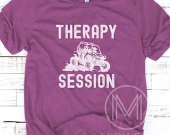 May include: A purple t-shirt with the words "THERAPY SESSION" in white, above and below a distressed graphic of an off-road vehicle. The shirt has short sleeves and a crew neck.