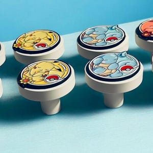 May include: A set of eight round white cabinet knobs featuring colorful circular illustrations of various creatures. Each knob displays a different character design, including green, yellow, blue, and orange figures, each accented with a small Poké Ball icon.