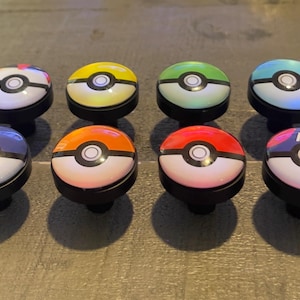 May include: Eight colorful cabinet knobs with a black base, each featuring a design reminiscent of a Poké Ball. The knobs have a white top with a central black circle and a colored band, including blue, yellow, green, and red.