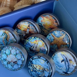 May include: A set of eight round, silver-toned, decorative drawer knobs with colourful ocean-themed images. The images include a lighthouse, a sailboat, a seahorse, a starfish, a turtle, a dolphin, and two different ocean sunset scenes.