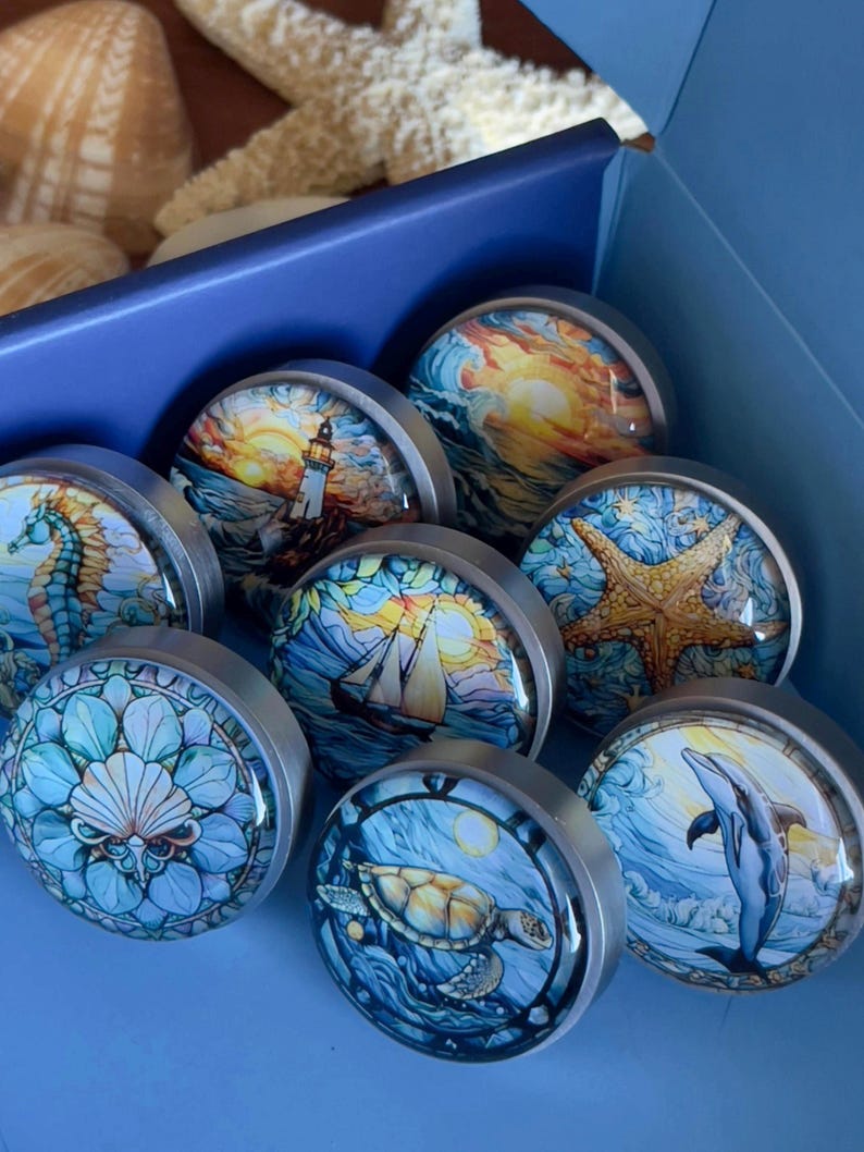 May include: A set of eight round, silver-toned, decorative drawer knobs with colourful ocean-themed images. The images include a lighthouse, a sailboat, a seahorse, a starfish, a turtle, a dolphin, and two different ocean sunset scenes.