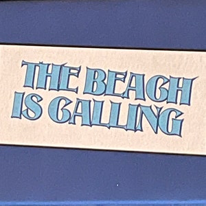Peut inclure: Une pancarte bleue et blanche avec le texte "The Beach is Calling" sur un fond beige clair.